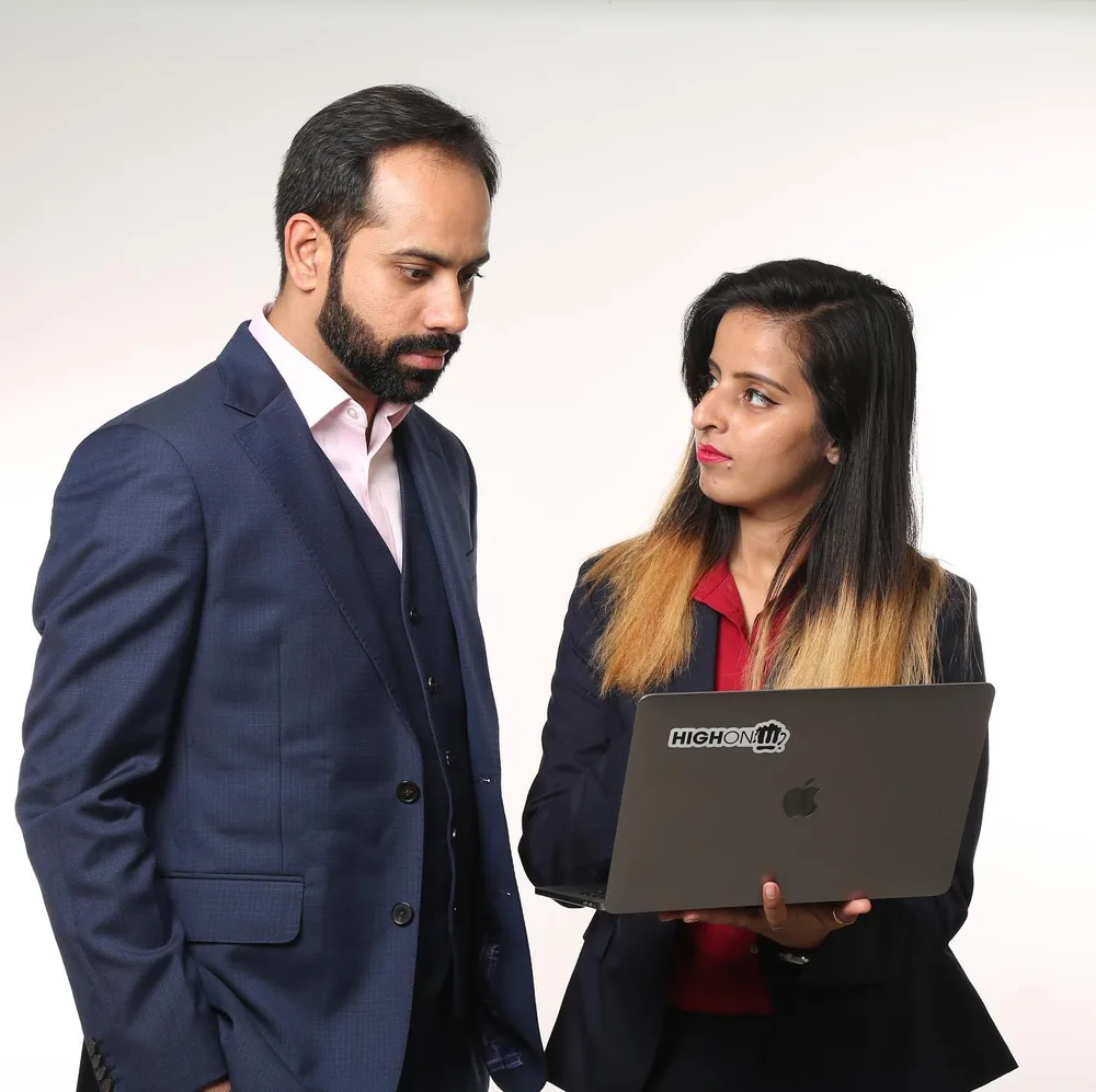 Studio photo: Gaurav in a navy suit and Roshni Dhal holding a HighonM laptop